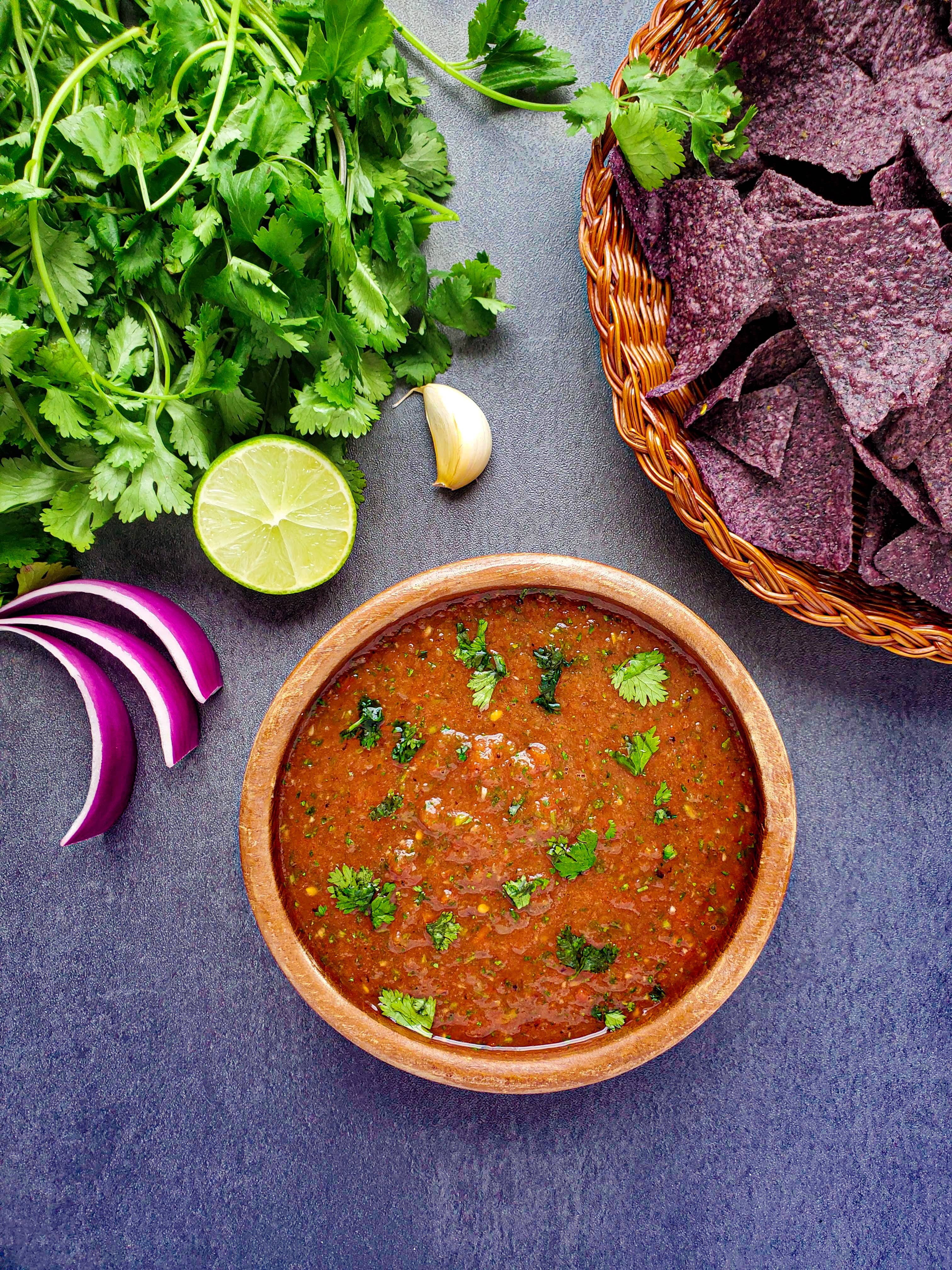 a bowl of mexican restaurant salsa with sliced red onion, a cut lime, a garlic clove, fresh cilantro and blue tortilla chips above the bowl of salsa