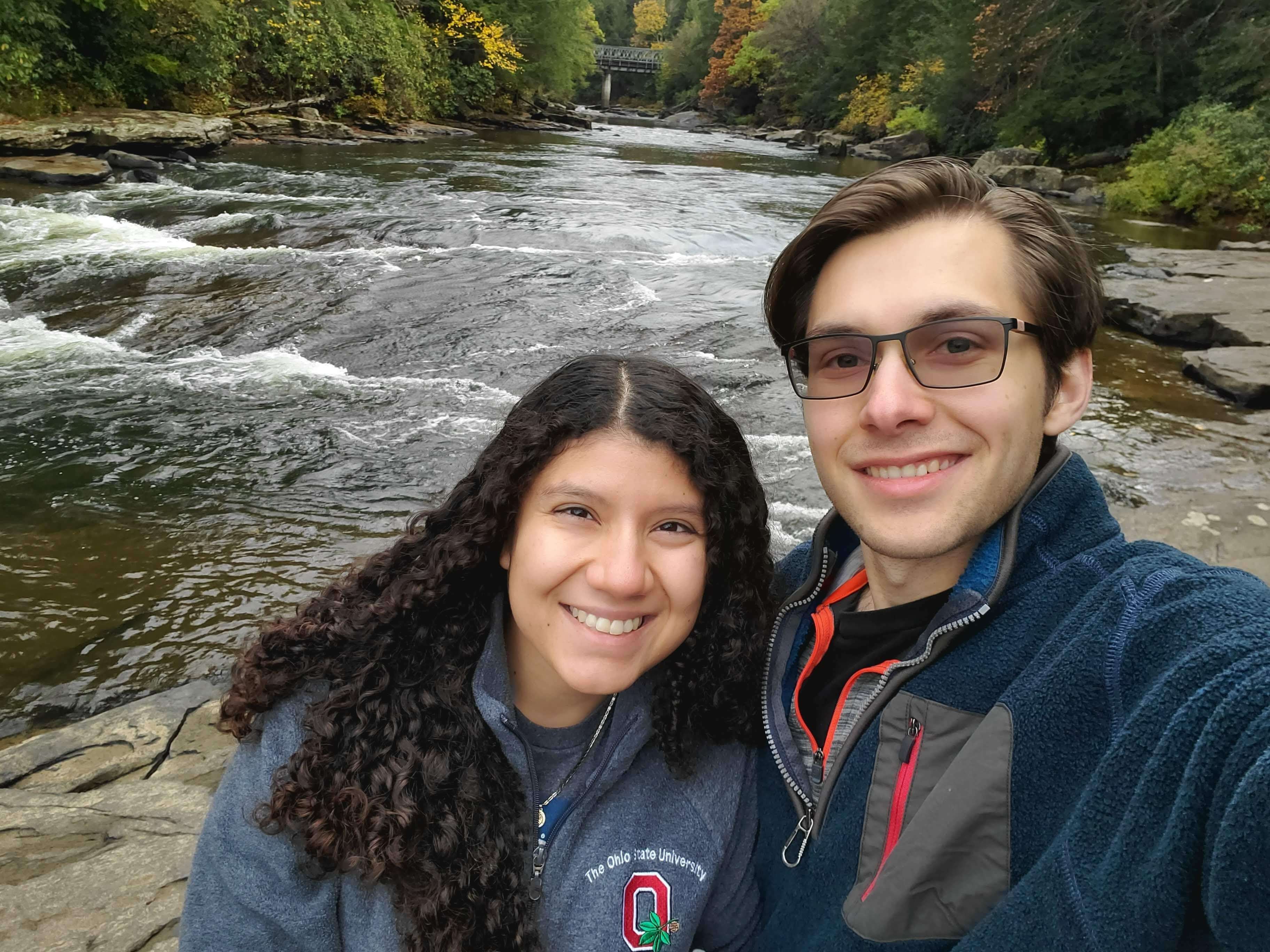a woman with dark curly hair and a man with short brown hair stand in front of a river smiling
