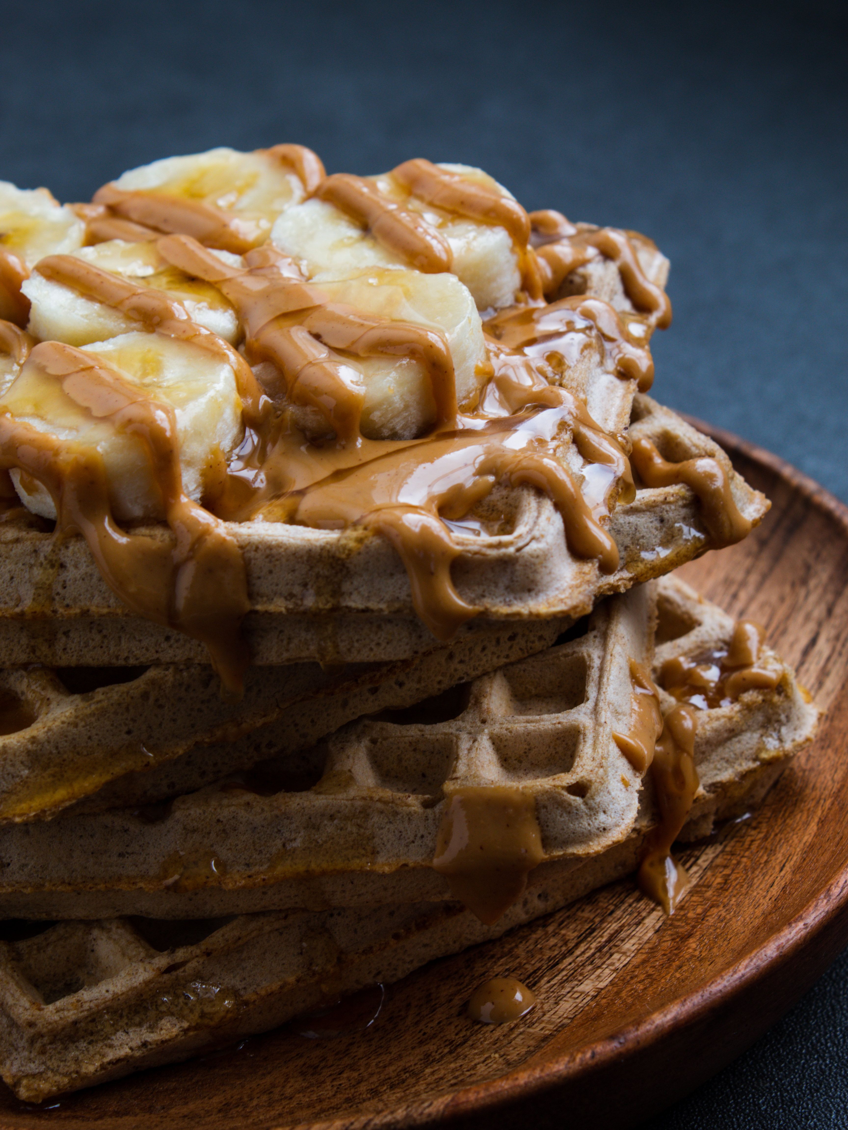 4 protein waffles stacked on top of a wooden plate with bananas, peanut butter and honey drizzled on top