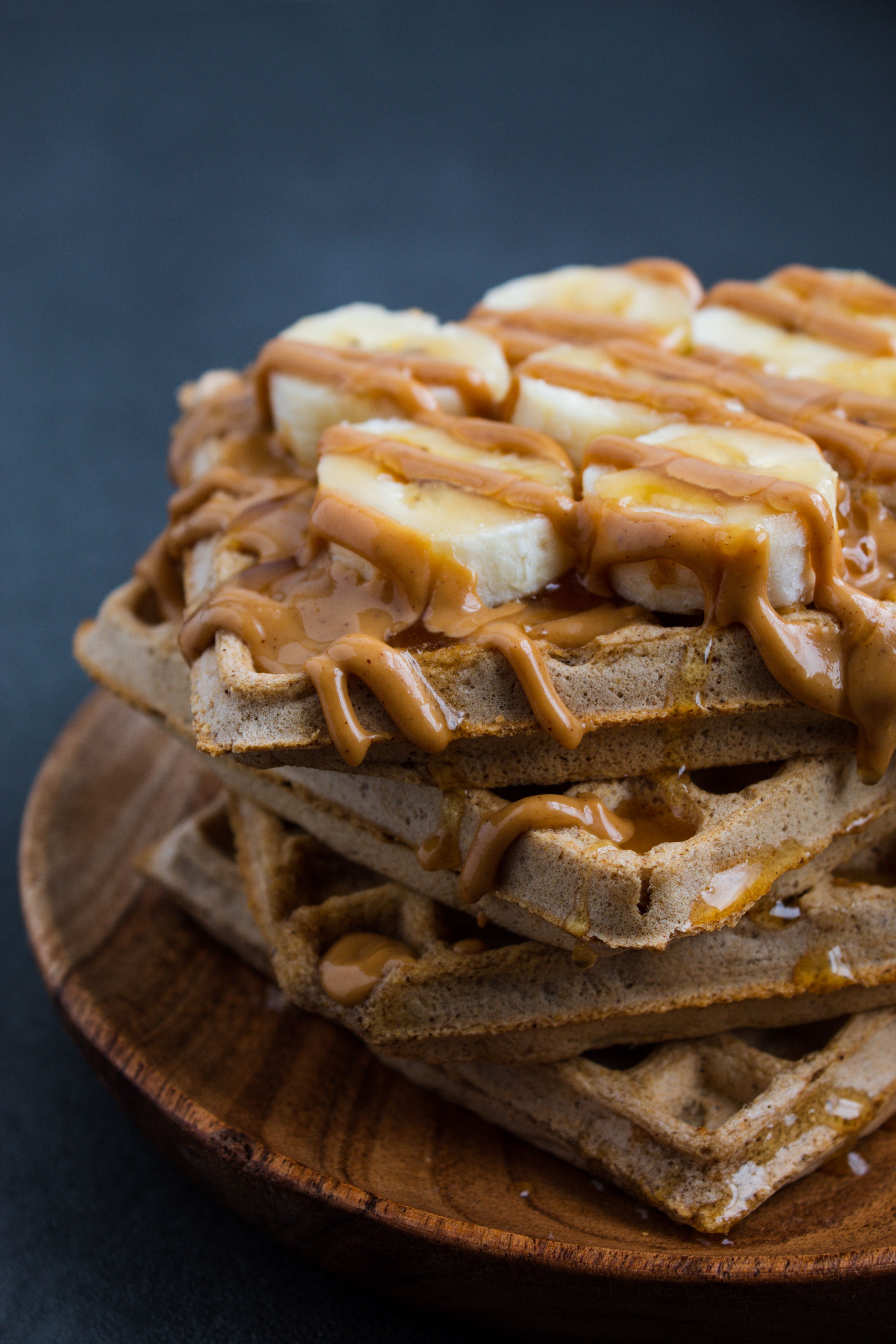 4 high protein waffles stacked on a wooden plate topped with banana slices and a drizzle of honey and peanut butter